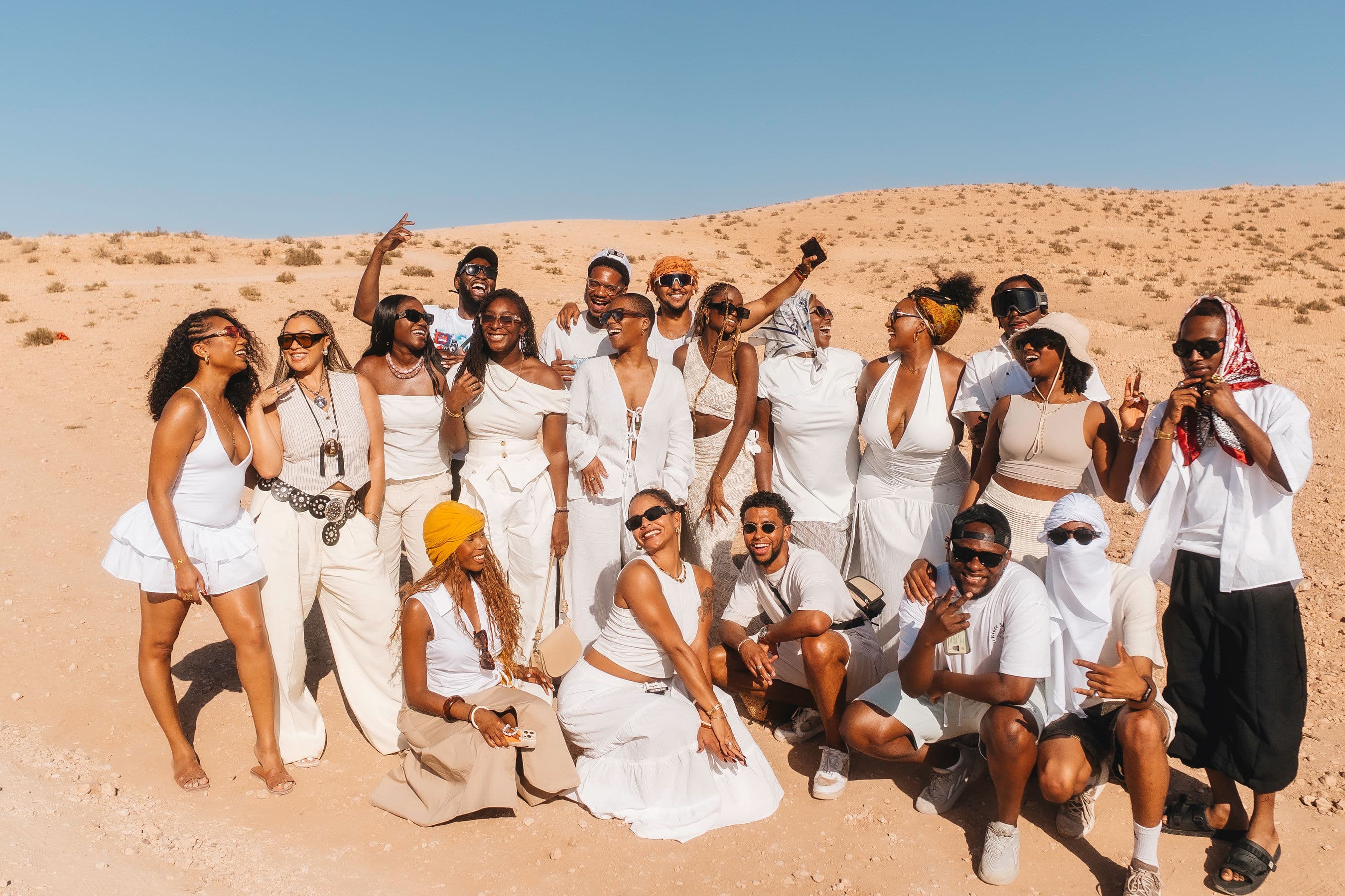 Large group of stylish people in white outfits posing together in a sunny desert landscape.